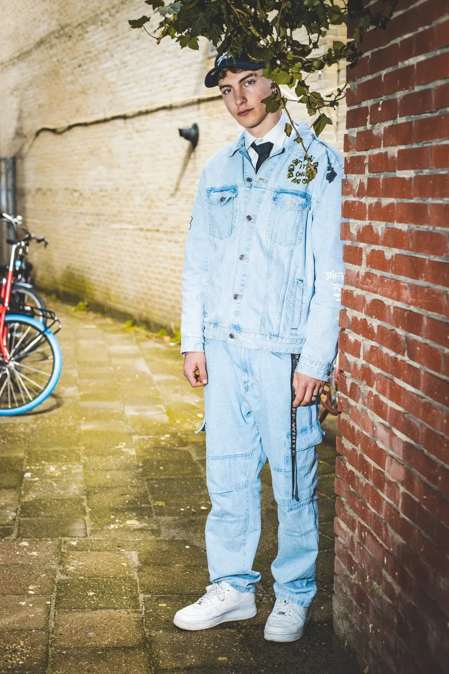 Jonge man in een lichtblauw denim pak en pet leunt tegen een bakstenen muur in een steegje